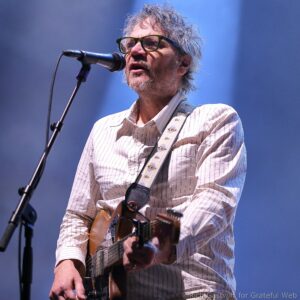 Male musician with gray hair and green glasses singing into a microphone while playing an acoustic guitar on stage.