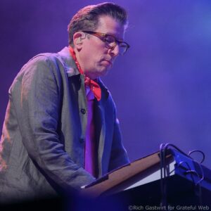 Musician wearing glasses and a red scarf plays a keyboard on a dimly lit stage with blue and purple lighting.