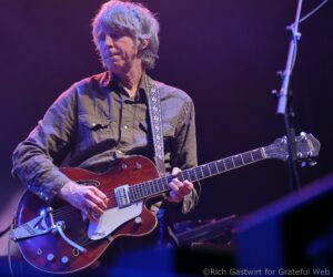 Male guitarist playing a sunburst electric guitar on stage under purple lighting with a patterned strap visible