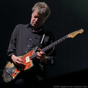 Guitarist playing a striped shirt on a dark stage, focus on orange-tinted guitar pickguard and his hands on the fretboard.