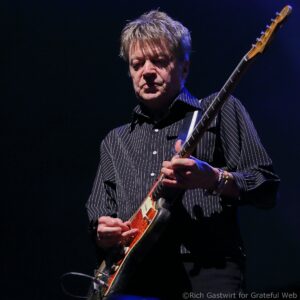 Male guitarist on stage playing a sunburst electric guitar, wearing a pinstriped shirt, illuminated by blue stage lighting.