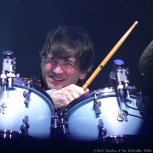Close-up of a drummer smiling with drumsticks raised, performing on a dark stage behind a drum kit.