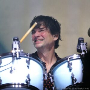 Close-up of a drummer's face with a drum kit visible in the foreground, smiling during performance.