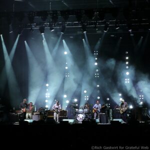 Live concert scene with multiple musicians on a dark stage, guitars and drums under bright spotlights as they perform.