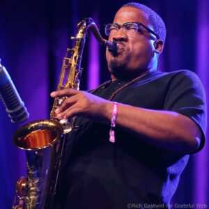 Close-up of a musician with glasses playing a gold saxophone on a purple-lit stage, focus on the face and instrument.