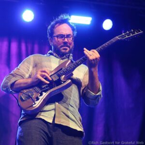 Guitarist with glasses and a beard, wearing a plaid shirt, playing an electric guitar on a blue-lit stage.