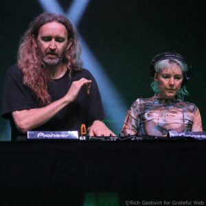 Two DJs perform on a dark stage: a bearded man with long hair speaks into a mic while a second person wearing headphones works the mixer.