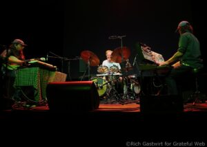 Three musicians on stage: guitarist left, drummer center, keyboardist right, under dark red-green lighting with their instruments visible.