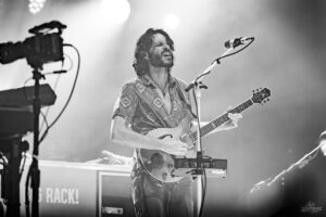 Male musician wearing a patterned shirt plays an electric guitar and sings into a microphone on a dimly lit stage with keyboards and gear in the background (black and white).