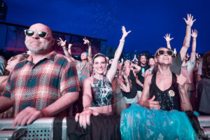 Crowd at an outdoor concert at night, front-row fans cheer with hands raised and smiles, barrier in the foreground.