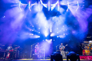 Band performs on a lit stage with blue-purple lights and fog, guitarist center, keyboardist left, drummer at right.