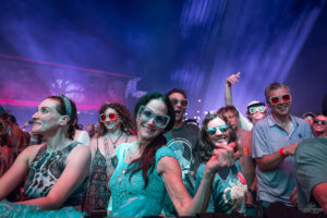 Crowd at a music festival wearing glittery sunglasses, dancing and smiling as blue and pink stage lights wash over them