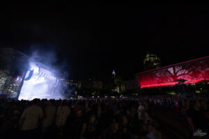 Nighttime outdoor concert with a smoke-filled, brightly lit stage on the left and a crowded audience; city skyline in the distance.