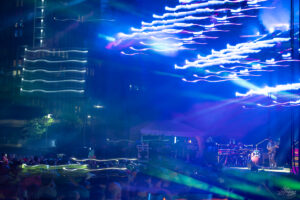 Outdoor concert at night: a band on a stage bathed in blue lights with colorful, streaking light trails and a crowd in the foreground.