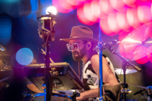Drummer wearing a brown hat and rhinestone sunglasses performing on a neon-lit concert stage.