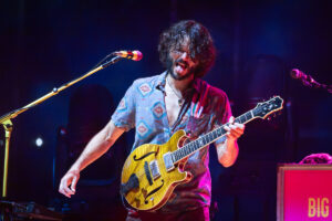 A guitarist on stage wearing a patterned shirt, playing a yellow electric guitar and sticking out his tongue in a lively performance.