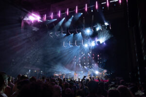 Concert stage at night with vibrant blue and pink lights, performers center stage, audience silhouettes, and fog effects creating a lively atmosphere