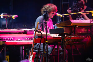 Musician with long hair singing into a mic while playing a red keyboard on stage, with a camera operator filming in the background