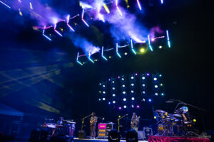 Band performs on a dark stage with bright vertical LED light bars, smoke effects, and a full drum kit on the right.