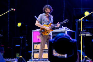 Male musician with a patterned short-sleeve shirt playing a wooden electric guitar on stage under blue lights during a live show, with gear in the background.
