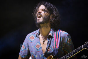 Male guitarist singing passionately on stage while playing an electric guitar, wearing a patterned blue shirt and a colorful strap over his shoulder.