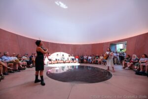 Violinist and guitarist perform inside a circular, open-air-like venue with an audience seated along curved pink walls.