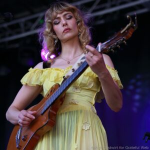 Female guitarist in a yellow off-shoulder dress playing a wooden guitar on stage, hands near the strings under stage lights.