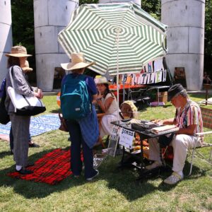 Group of people at an outdoor market under a large green-and-white striped umbrella, with a vendor at a table displaying items and signs nearby.