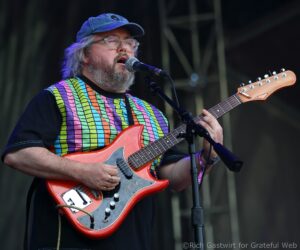Male guitarist with a gray beard wearing a blue cap and colorful plaid shirt, singing into a microphone while playing a red electric guitar on stage.