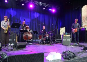 Four-piece band performing on a blue-curtained stage with a saxophonist, bassist, drummer, and guitarist under purple lights outdoors at a live show.