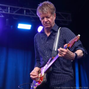 Male guitarist wearing a striped shirt playing a red electric guitar on stage under blue lights.
