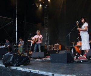 A live band performs on stage: guitarist center, keyboardist left, female singer on the right with a bass guitar, drums nearby, stage lights above.