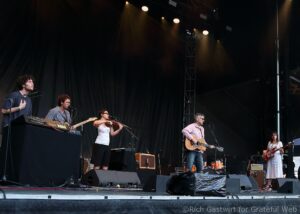 Stagewide view of a live band performing on an outdoor stage, with keyboard, guitar, violin, and microphone stands under warm yellow lights.