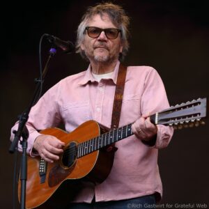 Male guitarist performing on stage, wearing a pink shirt and glasses, singing into a microphone.