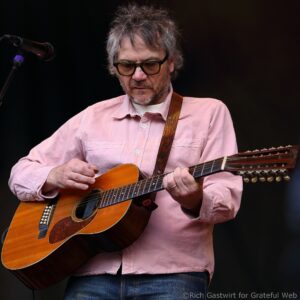 Male guitarist wearing a pink shirt, strumming an acoustic guitar on stage with a microphone nearby