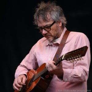 Man wearing a pink shirt plays a wooden acoustic guitar, close-up of hands strumming strings against a dark background