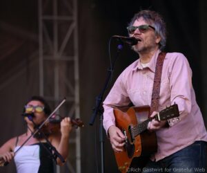 Male guitarist wearing a pale pink shirt and sunglasses performs on stage, singing into a microphone with a patterned strap on his guitar behind him is a violinist in a white top and glasses.