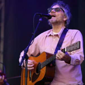 Male singer wearing glasses and a pink shirt sings into a microphone while playing an acoustic guitar on stage.