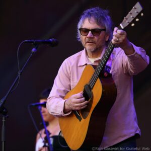 Male guitarist performing on stage, wearing sunglasses and a pink shirt, playing an acoustic guitar beside a mic stand.