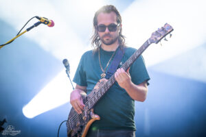 Male bassist wearing sunglasses and a teal shirt, playing a patterned bass on stage with bright backlighting.