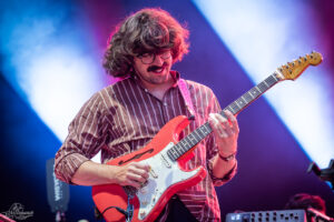 Male guitarist with wavy hair and a mustache, playing a red electric guitar on stage under blue and purple lights.