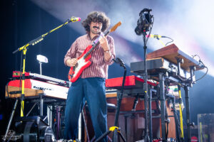 Male musician with wavy hair and mustache playing a red electric guitar on stage amidst keyboards and gear.