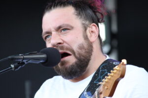 Close-up of a bearded man singing into a microphone while playing a guitar on stage, wearing a white shirt and a decorative strap.