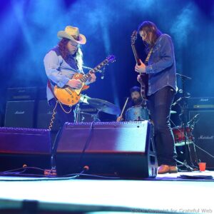 Two guitarists on a smoky blue-lit stage: one wearing a cowboy hat and denim, playing a sunburst electric guitar while the other in a denim jacket plays a darker guitar; drum kit and Marshall amps in the background.