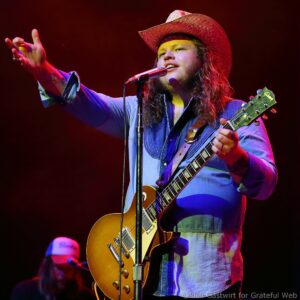 Male musician wearing a red cowboy hat plays a sunburst Les Paul-style guitar on stage, singing into a microphone against dark lighting.