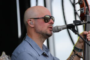 Bald man wearing sunglasses and an ear monitor, singing into a microphone while playing a guitar on stage.