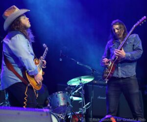 Two guitarists on a blue-lit stage: left musician wearing a wide-brimmed hat and denim jacket, right musician with long hair playing an electric guitar; a drum kit is visible in the background.