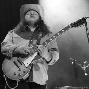 Guitarist in a hat and denim shirt playing a guitar on stage, close-up of the instrument and strings on his body.