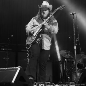 Guitarist wearing a cowboy hat and denim shirt, playing an electric guitar on stage with a microphone stand nearby.
