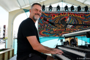Man in a black shirt playing a keyboard on an outdoor stage with a colorful, psychedelic banner in the background reading 'DEAD PANIC'.